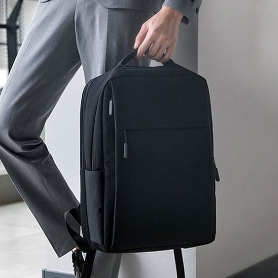 Homme en costume tenant un sac à dos ordinateur noir par la poignée, sac élégant et fonctionnel pour les déplacements professionnels.