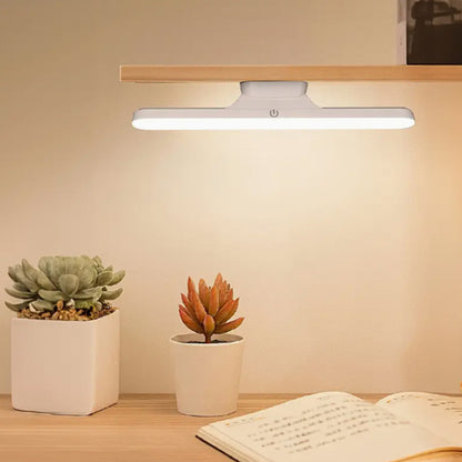 Lampe LED installée sous une étagère en bois, éclairant deux plantes et un livre
